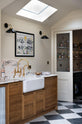 An oblique view of a grand and quaint Haberdasher’s kitchen features a sink and cooker run with a Paonazzo marble worktop and decorative vanity cut, a single Belfast sink, an aged brass Ionian tap, and a pantry cupboard. Beautifully decorated with tea towels, an artisan ceramic, a framed map illustration, black wall lights, a skylight, and a cloth lampshade, this kitchen creates a humble and functional space with a touch of elegance.