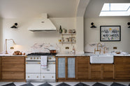 A grand and quaint Haberdasher’s kitchen features a sink and cooker run with a Paonazzo marble worktop and decorative vanity cut, a single Belfast sink, and an aged brass Ionian tap. Beautifully decorated with a white gas cooker with golden accents, a matching canopy, a deVOL grand tour desk lamp, a copper pan, artisan teapot, and dried ingredients in glass jars, this kitchen creates a humble and functional space with a touch of elegance.