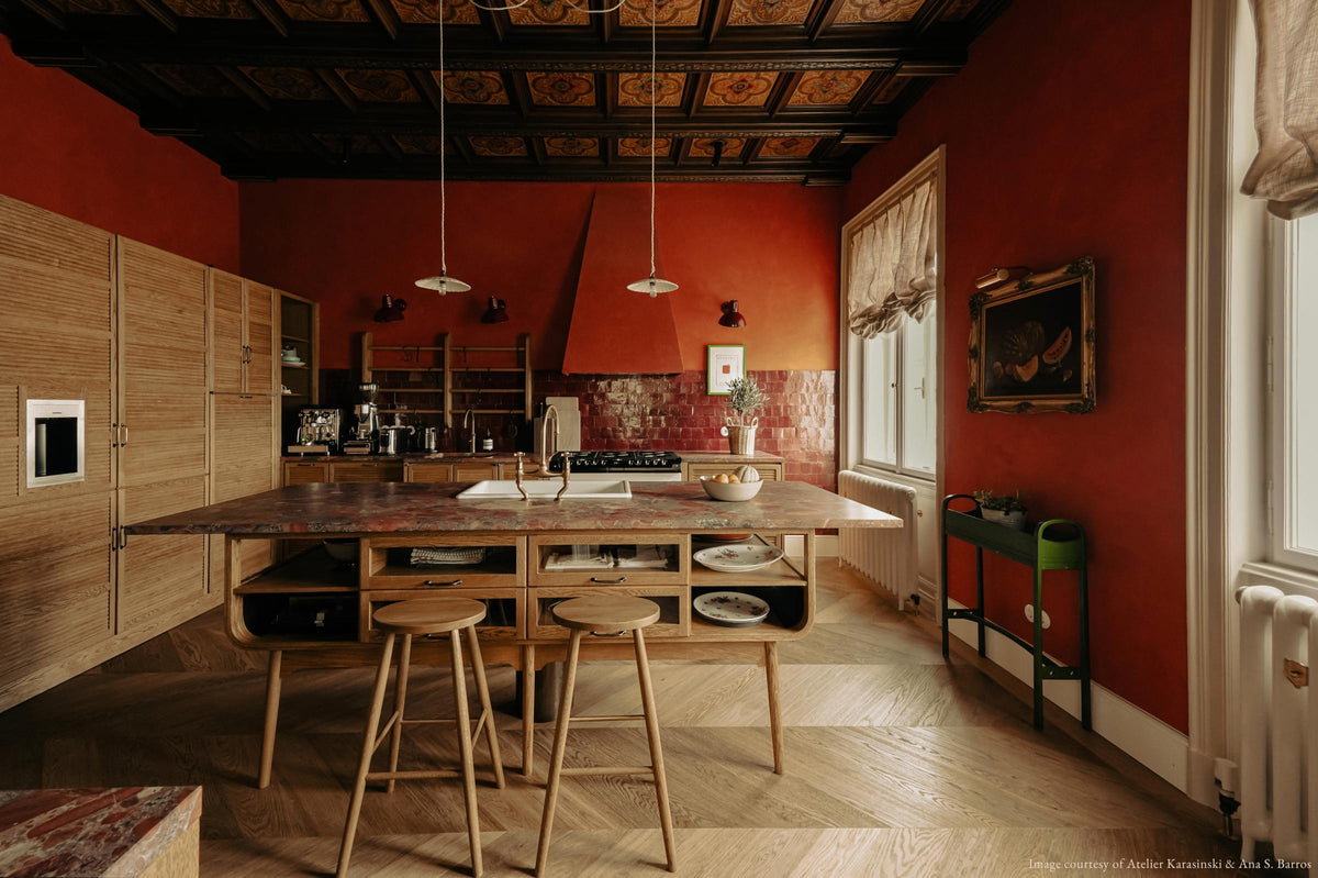 A warm and historical Haberdasher’s kitchen features a sink and cooker run with red Zellige splashback tiles, a kitchen island with an aged brass Ionian tap, a red marble worktop, and a bank of full-height cupboards. Beautifully decorated with decorative floral ceiling tiles, large narrow pleat porcelain lights, Helen stools, artisan ceramics, vintage items, and paintings, this kitchen creates a grand yet comfortable space.