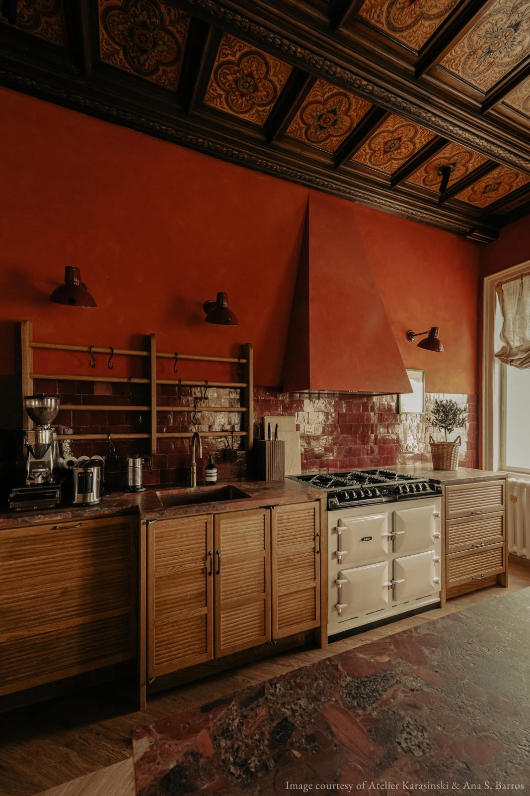 An oblique view of a warm and historical Haberdasher’s kitchen features a sink and cooker run with red Zellige splashback tiles and a kitchen island with a red marble worktop. Beautifully decorated with decorative floral ceiling tiles, artisan ceramics, vintage items, a silver coffee grinder, and a small house plant, this kitchen creates a grand yet comfortable space.