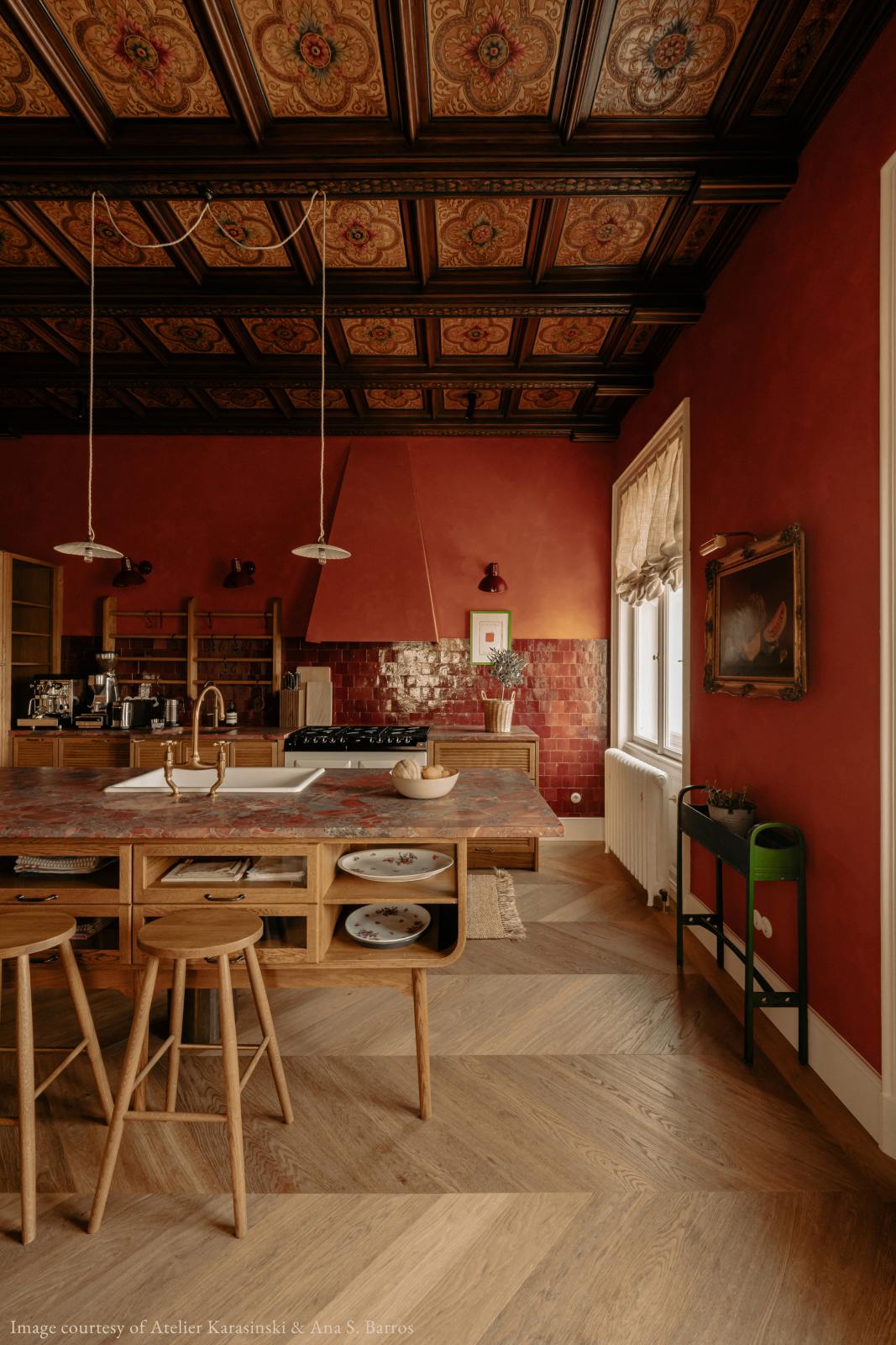 A warm and historical Haberdasher’s kitchen features a sink and cooker run with red Zellige splashback tiles, a kitchen island with an aged brass Ionian tap, and a red marble worktop. Beautifully decorated with decorative floral ceiling tiles, large narrow pleat porcelain lights, Helen stools, artisan ceramics, vintage items, and paintings, this kitchen creates a grand yet comfortable space.