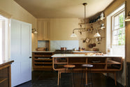 A cosy and sophisticated Haberdasher’s kitchen features a cooker run with a Carrara marble worktop, a small prep table, bespoke curved shelves, and muted yellow walls. Beautifully decorated with Creedy stools, a deVOL Heirloom gaselier light, Heirloom wall lights, aged brass hanging rails, copper pans, focaccia bread, vintage items, and artisan ceramics, this kitchen creates a bright and warm space.