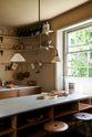 An oblique view of a cosy and sophisticated Haberdasher’s kitchen features a cooker run with a Carrara marble worktop, a small prep table, bespoke curved shelves, and muted yellow walls. Beautifully decorated with Creedy stools, a deVOL Heirloom gaselier light, aged brass hanging rails, copper pans, focaccia bread, vintage items, and artisan ceramics, this kitchen creates a bright and warm space.