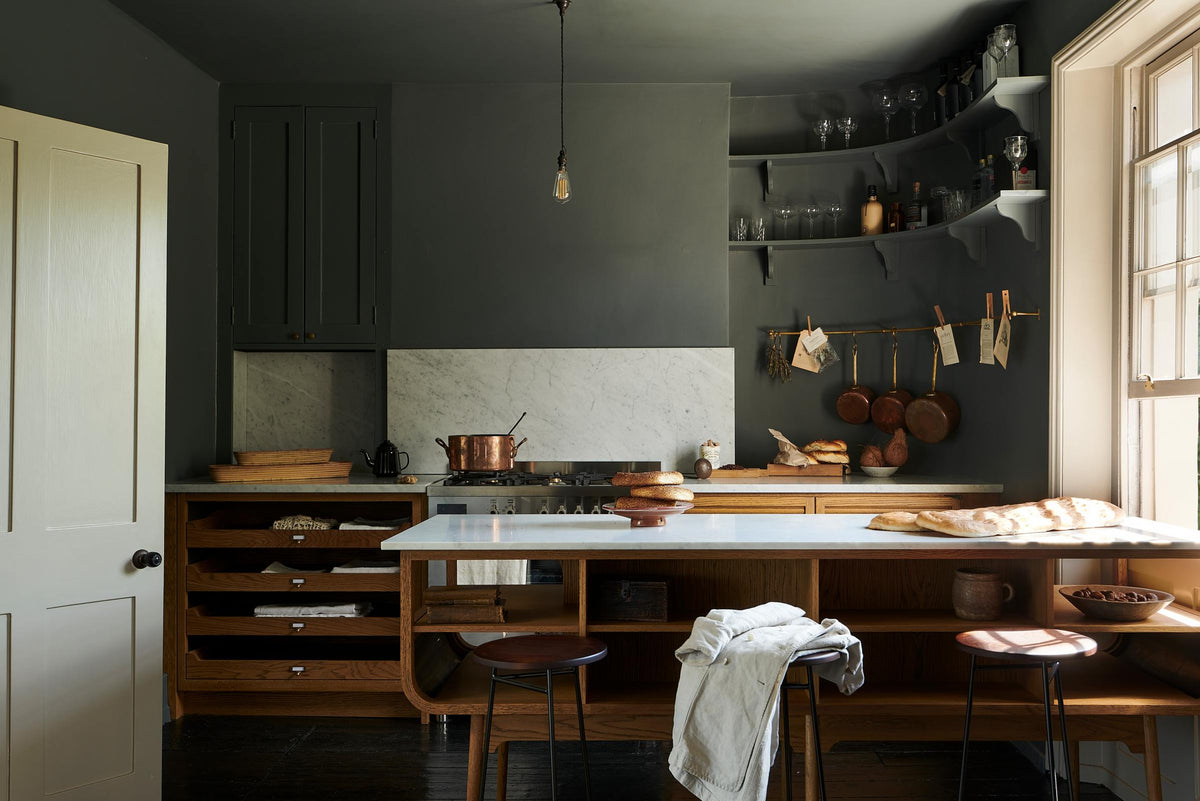 A cosy and sophisticated Haberdasher’s kitchen features a cooker run with a Carrara marble worktop, a small prep table, bespoke curved shelves, and dark charcoal walls. Beautifully decorated with stools, aged brass hanging rails, copper pans, fresh bread, vintage items, and artisan ceramics, this kitchen creates a cool yet warm and inviting space.