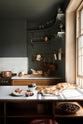 A closer look at a cosy and sophisticated Haberdasher’s kitchen features a cooker run with a Carrara marble worktop, a small prep table, bespoke curved shelves, and dark charcoal walls. Beautifully decorated with stools, aged brass hanging rails, copper pans, fresh bread, a bowl of chestnuts, figs, vintage items, and artisan ceramics, this kitchen creates a cool yet warm and inviting space.