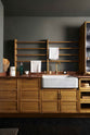 A closer look at a cosy and sophisticated Haberdasher’s kitchen features a sink run with a copper worktop, a single marble sink, glazed countertop cupboards, a freestanding pantry with glazed doors, and dark charcoal walls. Beautifully decorated with vintage furniture and items, wicker baskets, fresh bread, tea towels, and artisan ceramics, this kitchen creates a bright and warm space.