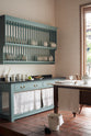 An oblique view of a beautiful and functional Trinity blue Heirloom kitchen features open cabinets, plate racks, and a showpiece Dairy table. Lovingly adorned with bone-white artisan ceramics, selected antiques, lace curtains, Gaselier lights, and earthy red terracotta square floor tiles that recreate Victorian kitchen furniture made to last a lifetime.