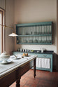 An oblique view through a doorway of a beautiful and functional Trinity blue Heirloom kitchen features open cabinets, plate racks, and a showpiece Dairy table. Lovingly adorned with bone-white artisan ceramics, selected antiques, lace curtains, Gaselier lights, and earthy red terracotta square floor tiles that recreate a Victorian kitchen made to last a lifetime.