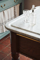 A close-up of the edge of a showpiece Heirloom Dairy table. Lovingly adorned with vintage crystal glasses and earthy red terracotta square floor tiles that recreate Victorian kitchen furniture made to last a lifetime.
