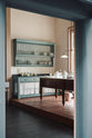 An oblique view through a door way of a beautiful and functional Trinity blue Heirloom kitchen features open cabinets, plate racks, and a showpiece Dairy table. Lovingly adorned with bone white artisan ceramics, selected antiques, lace curtains, Gaselier lights, and earthy red terracotta square floor tiles that recreate Victorian kitchen furniture that is made to last a lifetime.