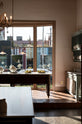 A dramatic photo of the sun shining through the large window of a beautiful and functional Trinity blue Heirloom kitchen features open cabinets, plate racks, and a showpiece Dairy table. Lovingly adorned with bone-white artisan ceramics, selected antiques, lace curtains, Gaselier lights, and earthy red terracotta square floor tiles that recreate a Victorian kitchen made to last a lifetime.