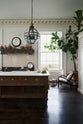A quiet corner of a simple yet grand Heirloom kitchen features a bespoke 3.5-metre-long dairy table with a single fluted Belfast sink, aged brass Mayan taps, a dishwasher, and a breakfast bar, creating a timeless and practical space. It is decorated with a vintage overmantle, artisan ceramics, glass and metal pendant lights, copper pans, and a large fiddle leaf fig plant.