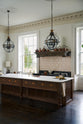 An oblique view of a simple yet grand Heirloom kitchen features a bespoke 3.5-metre-long dairy table with a single fluted Belfast sink, aged brass Mayan taps, a dishwasher, and a breakfast bar, creating a timeless and practical space. It is decorated with a vintage overmantle, artisan ceramics, glass and metal pendant lights, copper pans, and a large fiddle leaf fig plant.