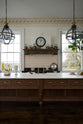 An oblique view of a simple yet grand Heirloom kitchen features a bespoke 3.5-metre-long dairy table with a single fluted Belfast sink, aged brass Mayan taps, a dishwasher, and a breakfast bar, creating a timeless and practical space. It is decorated with a vintage overmantle, artisan ceramics, glass and metal pendant lights, a plate of pears and lemons, and a large fiddle leaf fig plant.
