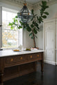 A quiet corner of a simple yet grand Heirloom kitchen features a bespoke 3.5-metre-long dairy table with a single fluted Belfast sink, aged brass Mayan taps, a dishwasher, and a breakfast bar, creating a timeless and practical space. It is decorated with a vintage overmantle, artisan ceramics, glass and metal pendant lights, Bentwood café chairs, and a golden-plated candelabra.