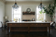 A simple yet grand Heirloom kitchen features a bespoke 3.5-metre-long dairy table with a single fluted Belfast sink, aged brass Mayan taps, a dishwasher, and a breakfast bar, creating a timeless and practical space. It is decorated with a vintage overmantle, artisan ceramics, glass and metal pendant lights, Bentwood café chairs, and a large fiddle leaf fig plant.