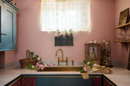 A small and practical Heirloom laundry room features a sink cabinet, a polished brass sink, an aged brass Ionian tap, a curtain rail with crest-patterned red curtains, wall cupboards, and shelves. Adorned with eclectic vintage items, pink peonies, and a small wicker basket, this space creates a romantic and functional atmosphere full of personality.