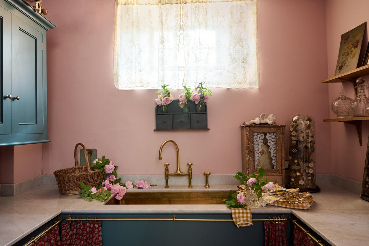 A small and practical Heirloom laundry room features a sink cabinet, a polished brass sink, an aged brass Ionian tap, a curtain rail with crest-patterned red curtains, wall cupboards, and shelves. Adorned with eclectic vintage items, pink peonies, and a small wicker basket, this space creates a romantic and functional atmosphere full of personality.