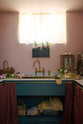 A small and practical Heirloom laundry room features a sink cabinet, a polished brass sink, an aged brass Ionian tap, a curtain rail with crest-patterned red curtains, and shelves. Adorned with eclectic vintage items, pink peonies, tea towels, and wicker baskets, this space creates a romantic and functional atmosphere full of personality.