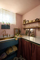 An oblique view of a small and practical Heirloom laundry room features a sink cabinet, a polished brass sink, an aged brass Ionian tap, a curtain rail with crest-patterned red curtains, and shelves. Adorned with eclectic vintage items, pink peonies, tea towels, glass containers, and wicker baskets, this space creates a romantic and functional atmosphere full of personality.