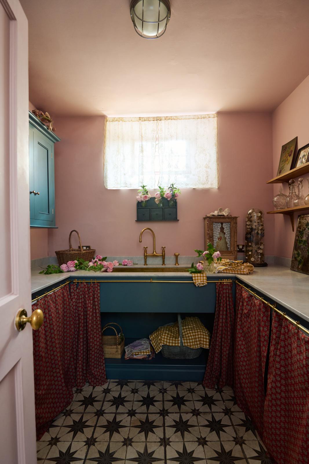 Past a blush pink door is a small and practical Heirloom laundry room featuring a sink cabinet, a polished brass sink, an aged brass Ionian tap, a curtain rail with crest-patterned red curtains, wall cupboards, and shelves. Adorned with eclectic vintage items, pink peonies, and a small wicker basket, this space creates a romantic and functional atmosphere full of personality.