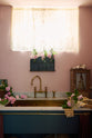 A closer look at a small and practical Heirloom laundry room features a sink cabinet, a polished brass sink, an aged brass Ionian tap, a curtain rail with crest-patterned red curtains, wall cupboards, and shelves. Adorned with eclectic vintage items, pink peonies, and a small wicker basket, this space creates a romantic and functional atmosphere full of personality.