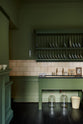 A quaint and stylish Wilkes green Heirloom kitchen features a sink cabinet with a café rail, white lace curtains, a polished brass sink, aged brass Mayan taps, and a large plate rack. Adorned with vintage photos, a cream bucket, simple ceramics, cream subway splashback tiles, and a golden-framed sliced green pumpkin, this kitchen creates a luxurious dining and social space.