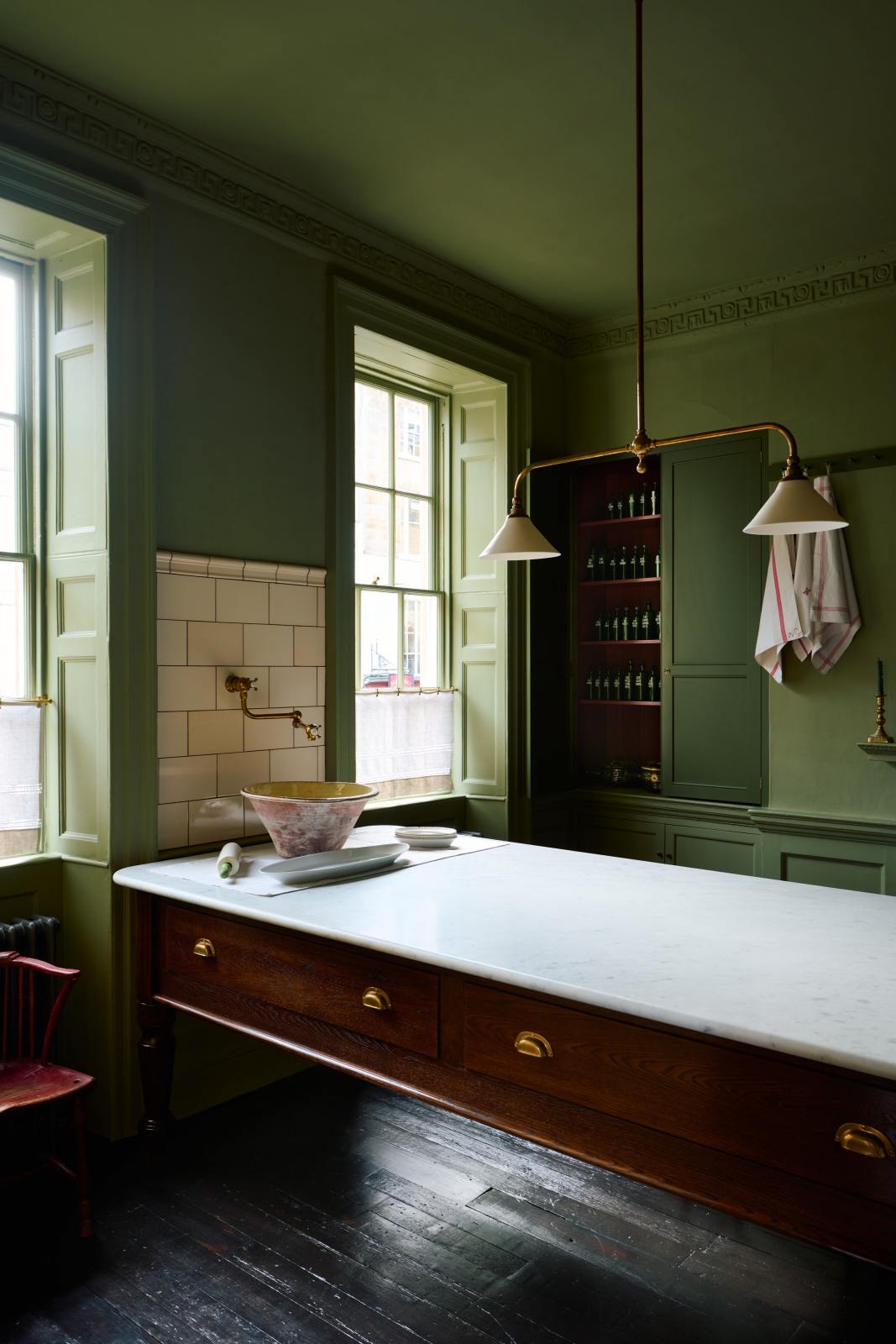 An oblique view of a quaint and stylish Wilkes green Heirloom kitchen features a large dairy table. Adorned with an aged brass pot filler, artisan ceramics, Heirloom gaselier lights, and cream subway splashback tiles, this kitchen creates a luxurious dining and social space.
