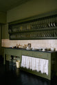An oblique view of a quaint and stylish Wilkes green Heirloom kitchen features a sink cabinet with a café rail, white lace curtains, a polished brass sink, aged brass Mayan taps, and a large plate rack. Adorned with vintage photos, a cream bucket, simple ceramics, cream subway splashback tiles, and a golden-framed sliced green pumpkin, this kitchen creates a luxurious dining and social space.