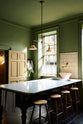 An oblique view of a quaint and stylish Wilkes green Heirloom kitchen features a large dairy table. Adorned with an aged brass pot filler, artisan ceramics, Heirloom gaselier lights, Helen stools, and cream subway splashback tiles, this kitchen creates a luxurious dining and social space.