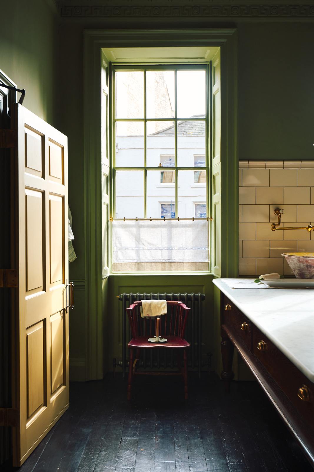 A quiet corner of a quaint and stylish Wilkes green Heirloom kitchen features a large dairy table, aged brass pot filler tap, and cream subway splashback tiles. Adorned with a café rail, a lace curtain, vintage coat hooks, and a small wooden chair, this kitchen creates a luxurious dining and social space.