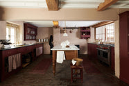 A refectory-red Heirloom kitchen features a sink cabinet with a polished brass sink, aged brass Mayan taps, café curtain rails, a red and white striped curtain, a dairy table, a cooker run, a vegetable box, and a plate rack. Beautifully decorated with Heirloom gaselier lights, a pot hanger, a vintage scale, artisan ceramics, and dried ingredients, this kitchen offers a modern twist on vintage furniture, creating a classic range designed to last.