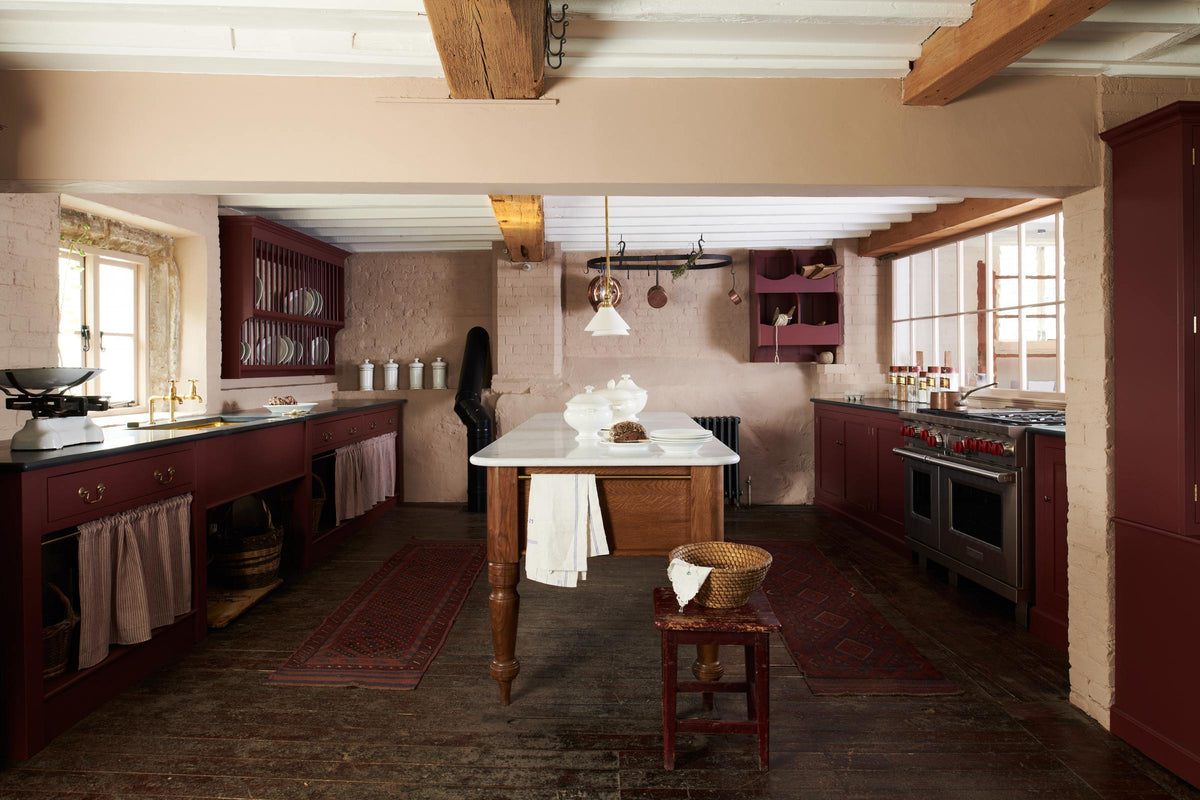 A refectory-red Heirloom kitchen features a sink cabinet with a polished brass sink, aged brass Mayan taps, café curtain rails, a red and white striped curtain, a dairy table, a cooker run, a vegetable box, and a plate rack. Beautifully decorated with Heirloom gaselier lights, a pot hanger, a vintage scale, artisan ceramics, and dried ingredients, this kitchen offers a modern twist on vintage furniture, creating a classic range designed to last.