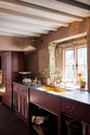 A closer view of a refectory-red Heirloom kitchen’s sink cabinet reveals a polished brass sink, aged brass Mayan taps, café curtain rails, a red and white striped curtain, and a pantry cupboard. Beautifully decorated with an Heirloom task light, a vintage scale, artisan ceramics, and house plants, this kitchen offers a modern twist on vintage furniture, creating a classic range designed to last.