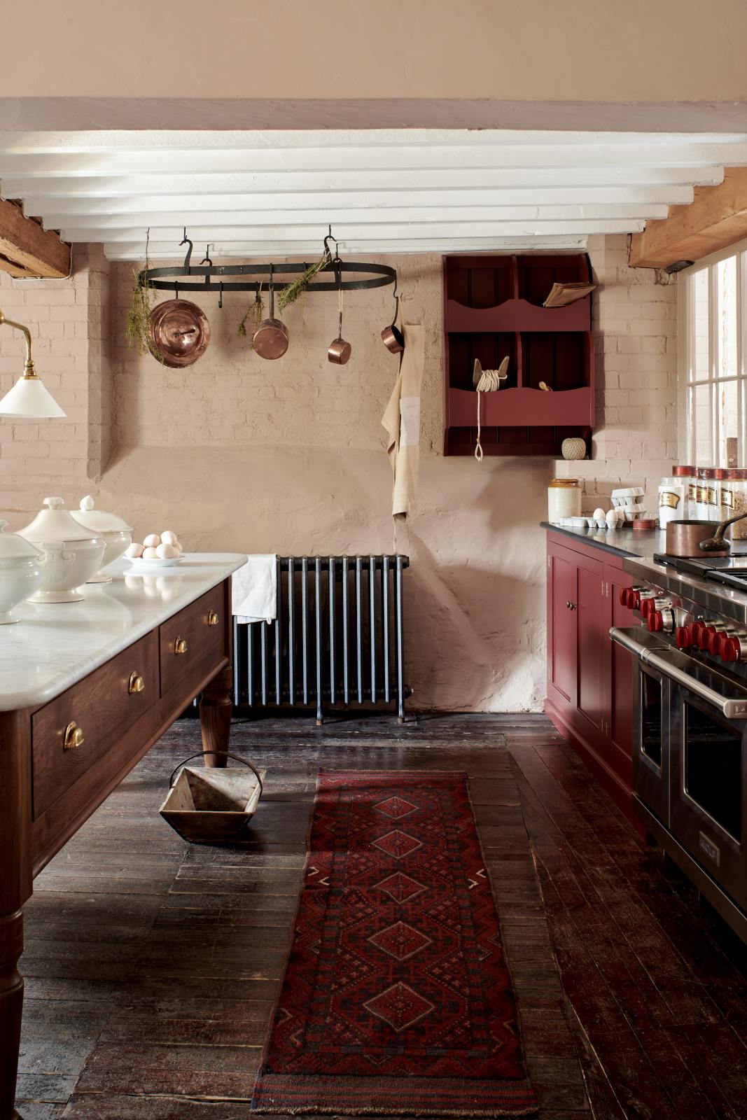 A refectory-red Heirloom kitchen features a dairy table, a cooker run, and a vegetable box. Beautifully decorated with Heirloom gaselier lights, a pot hanger, copper pans, artisan ceramics, and dried ingredients, this kitchen offers a modern twist on vintage furniture, creating a classic range designed to last.