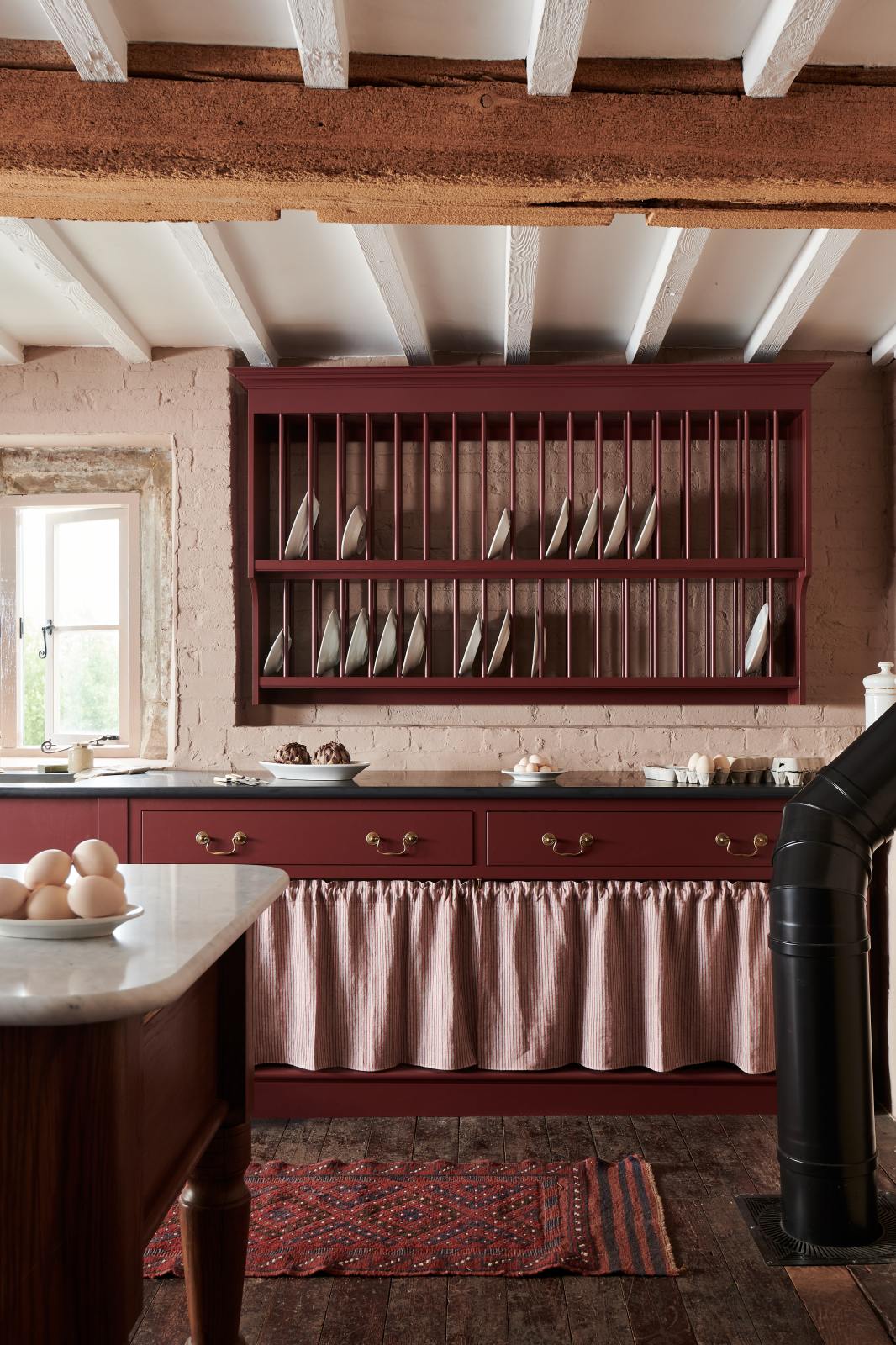 A refectory-red Heirloom kitchen’s sink cabinet reveals a polished brass sink, aged brass Mayan taps, café curtain rails, a red and white striped curtain, a plate rack, and an Heirloom dairy table. Beautifully decorated with artisan ceramics and plates of fresh eggs, this kitchen offers a modern twist on vintage furniture, creating a classic range designed to last.