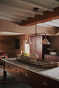 An oblique view of a refectory-red Heirloom kitchen features a sink cabinet with café curtain rails, a red and white striped curtain, a dairy table, and a pantry cupboard. Beautifully decorated with Heirloom gaselier lights, a vintage scale, artisan ceramics, and a large woven placemat, this kitchen offers a modern twist on vintage furniture, creating a classic range designed to last.
