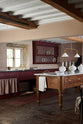 An oblique view of a refectory-red Heirloom kitchen features a sink cabinet with a polished brass sink, aged brass Mayan taps, café curtain rails, a red and white striped curtain, a dairy table, and a plate rack. Beautifully decorated with Heirloom gaselier lights, artisan ceramics, and a loaf of bread, this kitchen offers a modern twist on vintage furniture, creating a classic range designed to last.