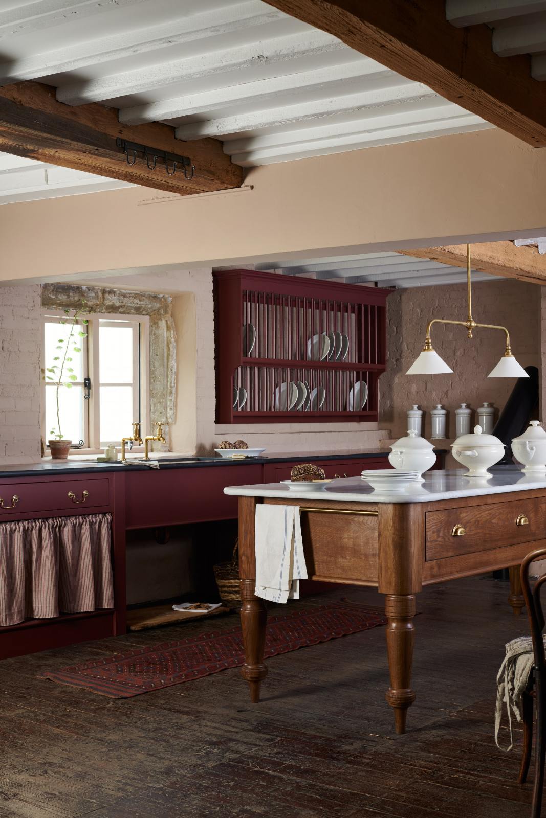 An oblique view of a refectory-red Heirloom kitchen features a sink cabinet with a polished brass sink, aged brass Mayan taps, café curtain rails, a red and white striped curtain, a dairy table, and a plate rack. Beautifully decorated with Heirloom gaselier lights, artisan ceramics, and a loaf of bread, this kitchen offers a modern twist on vintage furniture, creating a classic range designed to last.