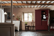 A quiet corner of a refectory-red Heirloom kitchen features a pantry cupboard. Beautifully decorated with vintage furniture, house plants, a creamware pendant light, and an aged brass café curtain rail, this kitchen offers a modern twist on vintage furniture, creating a classic space designed to last.