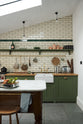 A quiet corner of a quaint and modern Wilkes green Shaker kitchen features a cooker and sink run with a rustic oak worktop, a long shaker shelf, and a Dairy table with a breakfast nook. Adorned with a long hanging rail, signature ceramics, brass cups, and a large wall of metro tiles it embodies a playful industrial and practical design.