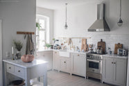 A humble Mushroom colour shaker kitchen with an all-in-one L-shaped sink and cooker run, and a small rolling kitchen island with Carrara marble worktop. A single Belfast sink is complemented by an aged brass Ionian tap, and an aged brass hanging rail adds a striking element against the grey and neutral tones of this charming kitchen.
