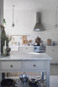 A closer look at the small, rolling kitchen island featuring a Carrara marble worktop. It overlooks the rest of the shaker kitchen, which includes an all-in-one L-shaped sink and a cooker run with a single Belfast sink. This setup is complemented by an aged brass Ionian tap, an aged brass hanging rail, and white crackle metro tiles.