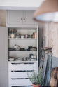 A close-up of an open floor-to-ceiling pantry cupboard in a Mushroom colour, storing lovingly collected ceramics and jar-sealed dry ingredients, surrounded by vintage furniture that fits perfectly at home.