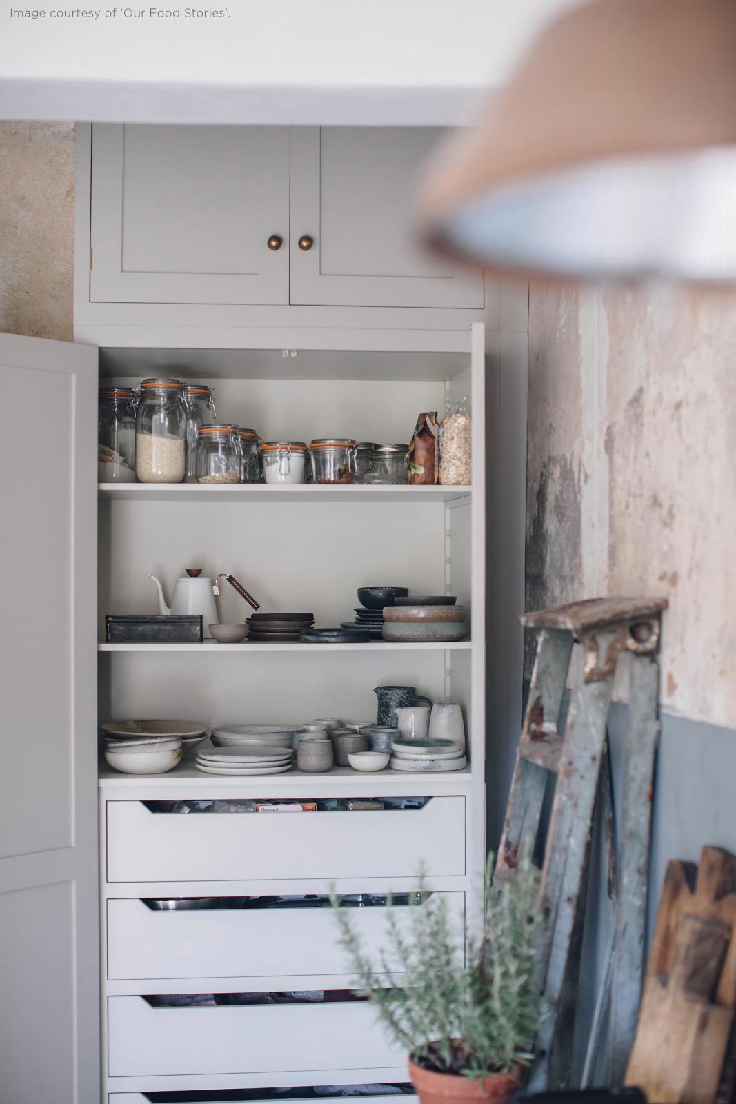 A close-up of an open floor-to-ceiling pantry cupboard in a Mushroom colour, storing lovingly collected ceramics and jar-sealed dry ingredients, surrounded by vintage furniture that fits perfectly at home.