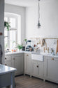 An angled corner of the humble Mushroom colour shaker kitchen with an all-in-one L-shaped sink and cooker run, and a small rolling kitchen island with Carrara marble worktop. A single Belfast sink is complemented by an aged brass Ionian tap, and an aged brass hanging rail adds a striking element against the grey and neutral tones of this charming kitchen.