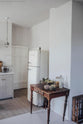 In a corner of the Mushroom-coloured shaker kitchen, there is an L-shaped sink and cooking area with a Carrara marble worktop. The large Smeg fridge stands out next to a vintage wooden desk adorned with collected items and fresh produce.