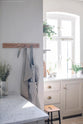 In the corner of a mushroom-coloured shaker kitchen, an L-shaped sink and cooker are featured alongside a small rolling island with a Carrara marble worktop. Wooden hanging racks hold two aprons and golden-handled scissors for added functionality.