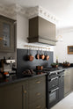 An oblique view of a murky-green galley shaker kitchen, featuring a cooker run with a black granite worktop and splashback, a shaker canopy, a glazed wall cupboard, and an aged brass hanging rail. Beautifully curated with a vintage gold-framed painting, artisan ceramics, fresh produce, houseplants, and copper pans, it creates a chic, understated, practical galley kitchen.