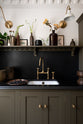 A closer look at the murky-green galley shaker kitchen, featuring a sink run with a black granite worktop and splashback, an undermounted sink, an aged brass ionian tap, and a shaker shelf with pegs. Beautifully curated with two brass wall lights, artisan ceramics, fresh houseplants, framed vintage illustrations, and a brass plate of blackberries, it creates a chic, understated, and practical galley kitchen.